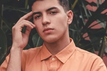 A model in an orange short-sleeved shirt resting a hand against their temple, framed by tropical foliage.